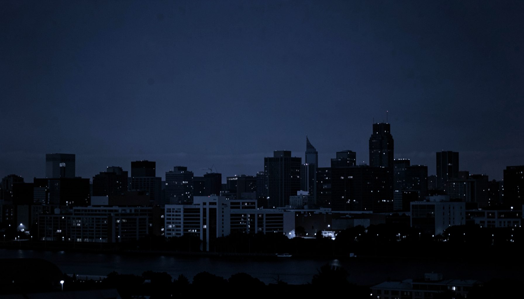 Muted monochrome photograph of the Minneapolis skyline at dusk, deep blue tones blending into a dark navy backdrop